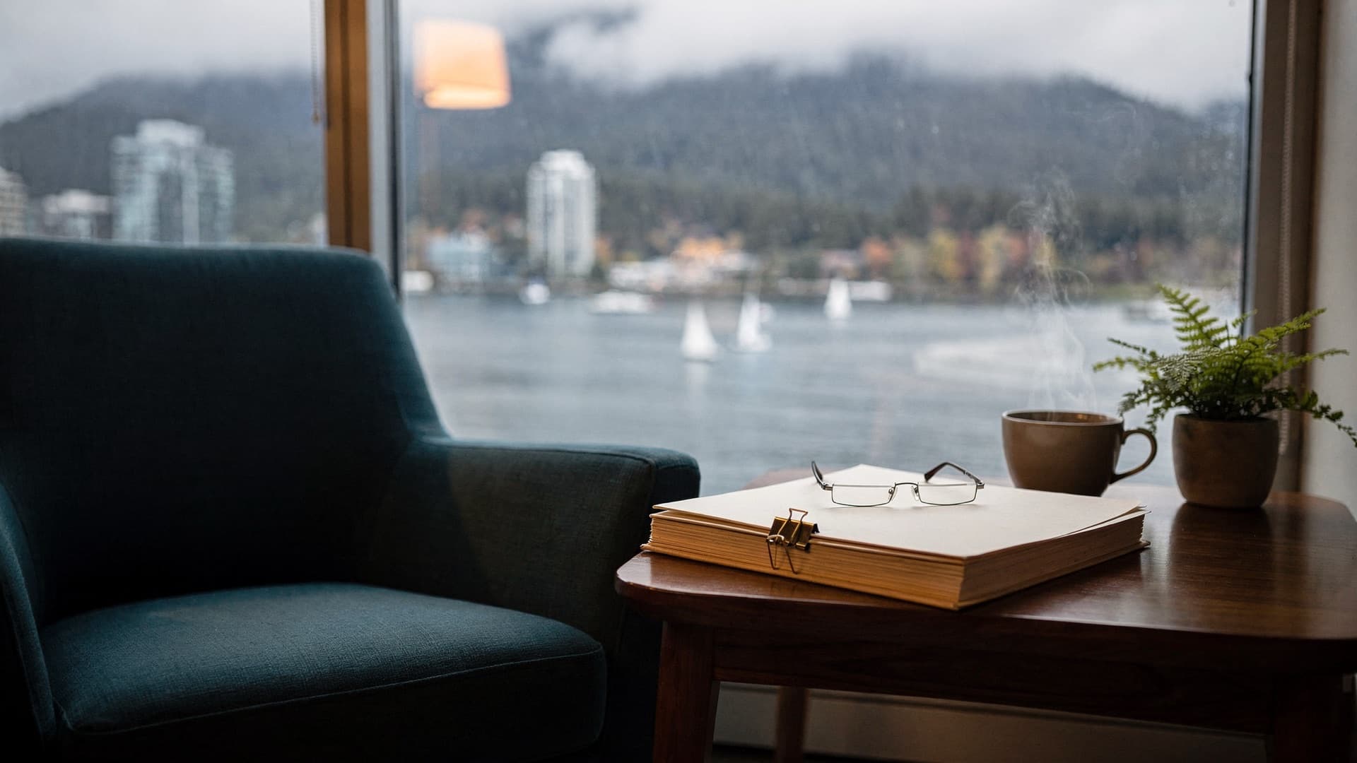 A teal armchair beside a side table with blank folders, reading glasses, a cup of tea and a fern, by a window overlooking a misty Vancouver harbour with sailboats and forested mountains
